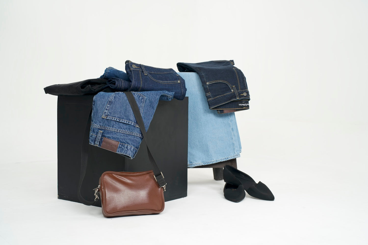 Black box with folded jeans, a brown leather wallet, and black shoes on a white background