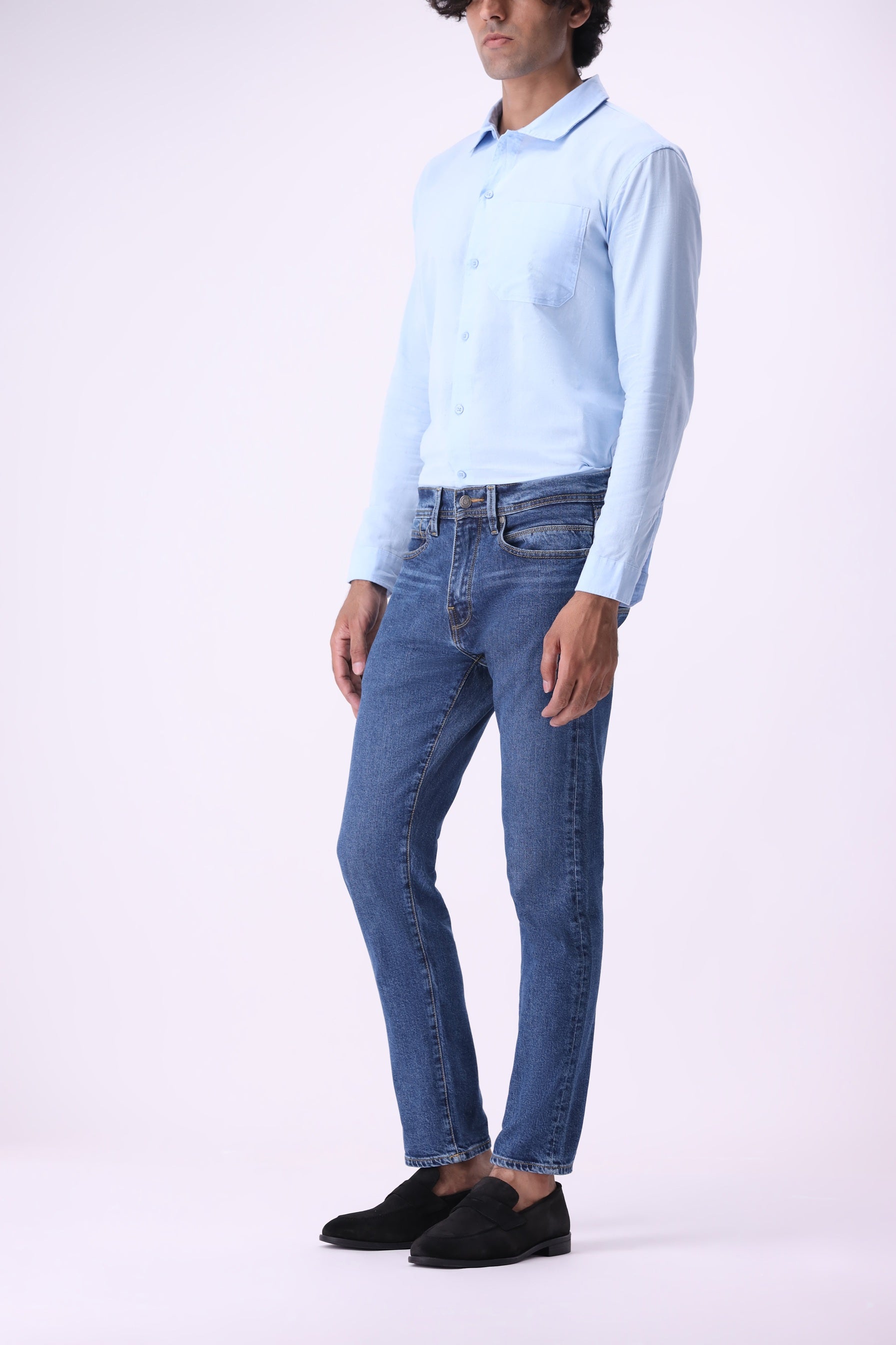 Side angle of men’s Baloo slim fit medium indigo wash jeans with light blue shirt on plain white background