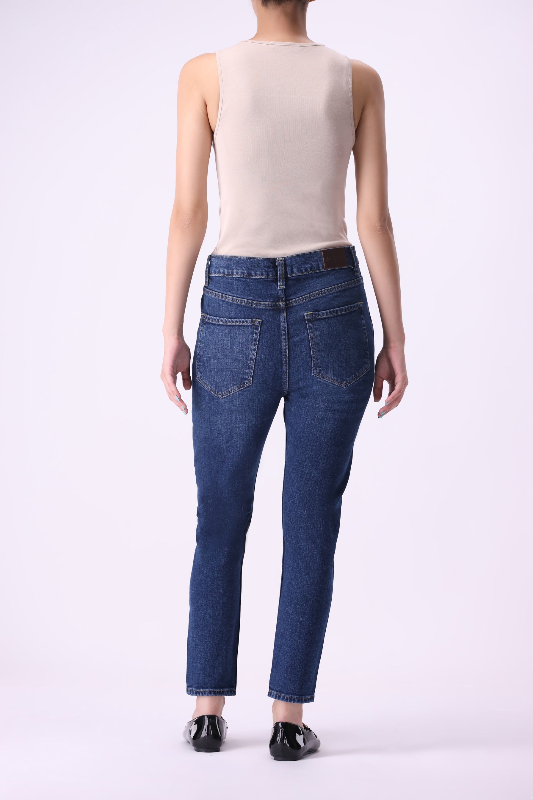 Full back view of women’s Beryoza jeans in medium dark indigo wash, slim tapered fit, paired with a beige tank top.